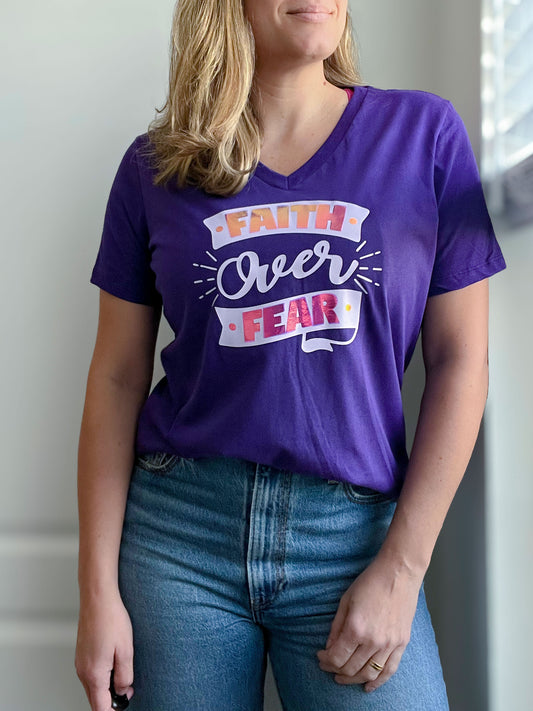 A woman wearing a deep purple V-neck T-shirt featuring the phrase “Faith Over Fear” in a bold and decorative design. The text incorporates a mix of light purple and pink holographic elements, making it stand out with a reflective effect. She pairs the shirt with classic blue jeans, creating a stylish and faith-inspired look. Part of Spark of Faith’s handmade apparel collection.