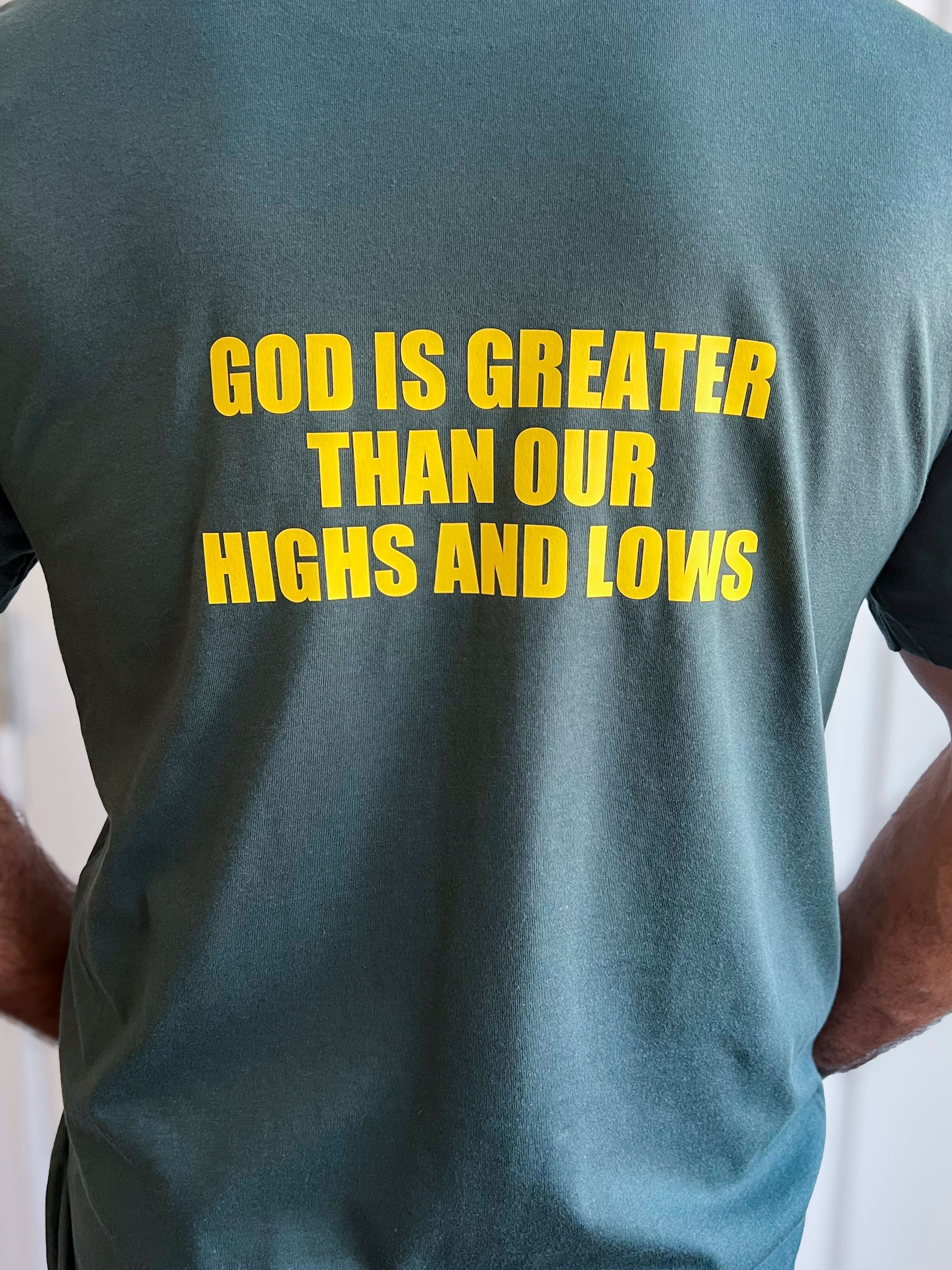 Back of a forest green T-shirt with large yellow text reading ‘GOD IS GREATER THAN OUR HIGHS AND LOWS.