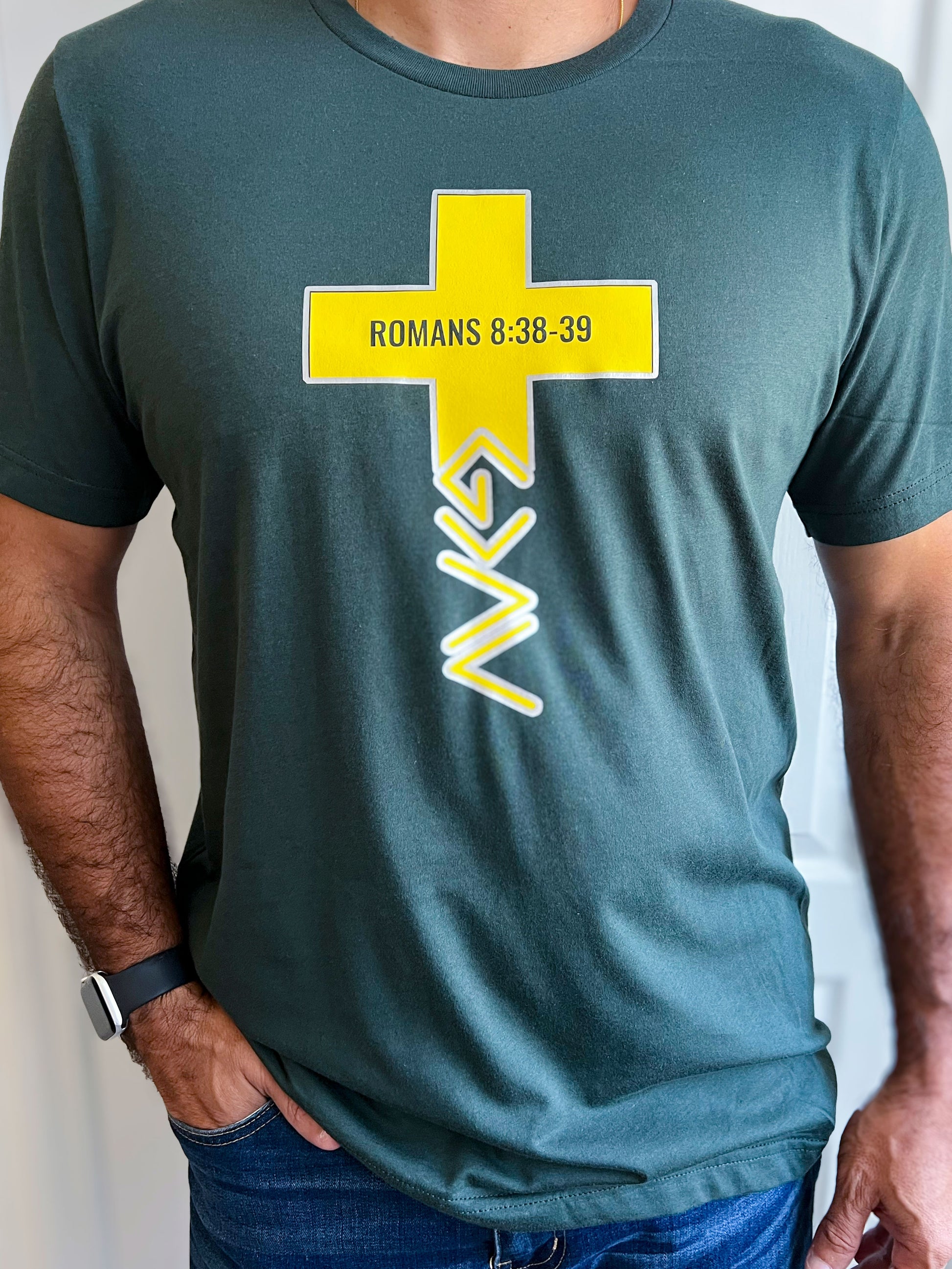 Man wearing a forest green T-shirt featuring a bold yellow cross with ‘ROMANS 8:38-39’ printed inside. Below the cross is a stylized symbol representing ‘God is greater than our highs and lows.’ Styled with blue jeans.