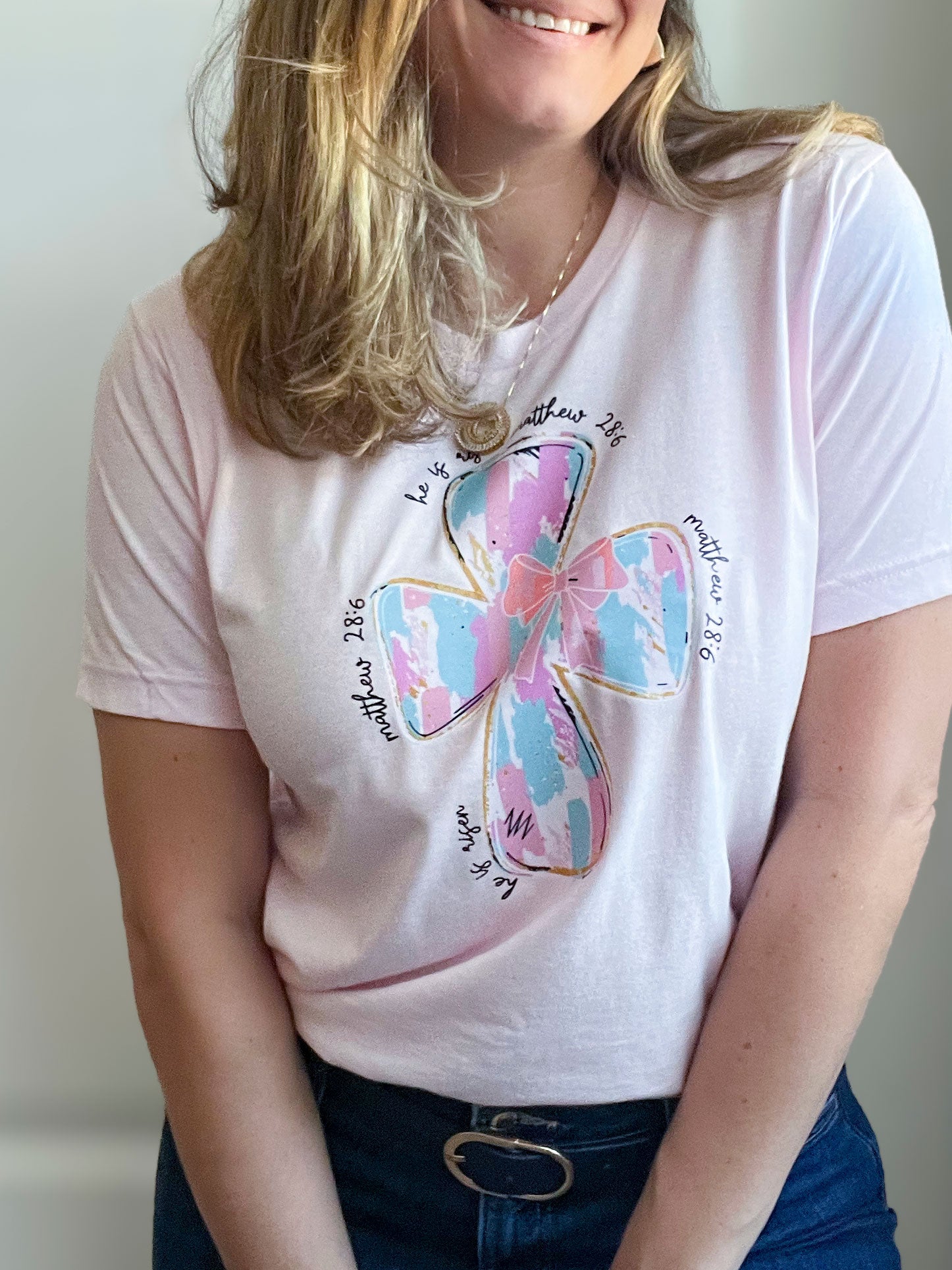 Woman wearing a light pink T-shirt featuring a large pastel cross design in shades of blue, pink, and white with gold outlines. Surrounding the cross, the text reads ‘He is risen’ and ‘Matthew 28:6’ in a handwritten-style font. Styled with dark blue jeans, a blue belt with a gold buckle, and a delicate gold necklace.