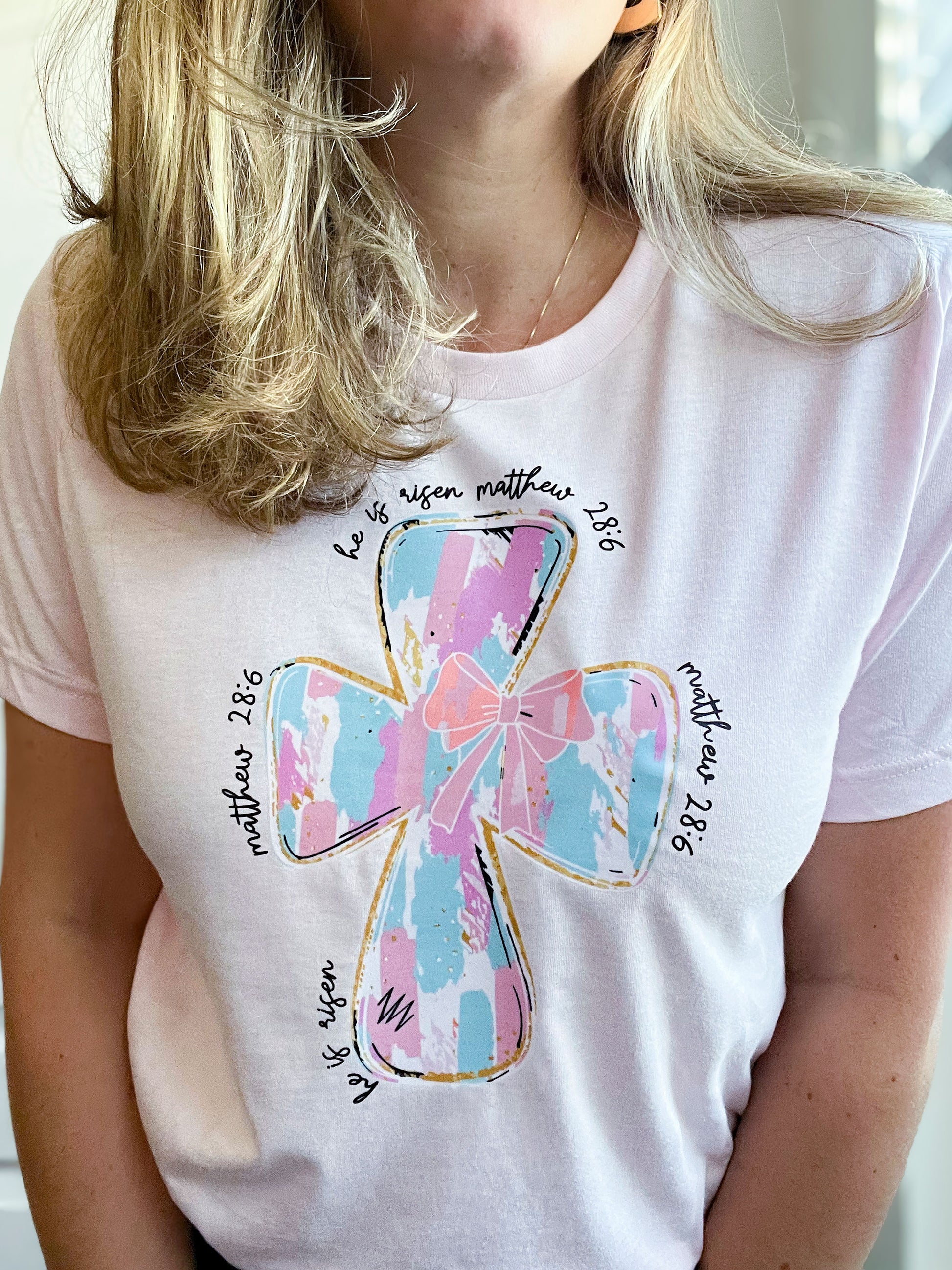 Woman wearing a light pink T-shirt featuring a large pastel cross design in shades of blue, pink, and white with gold outlines. Surrounding the cross, the text reads ‘He is risen’ and ‘Matthew 28:6’ in a handwritten-style font. 