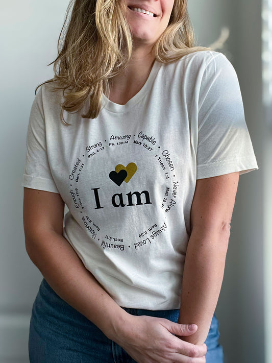 A woman wearing a vintage white T-shirt with the phrase “I am” in bold black letters, accented by two hearts—one black and one gold glitter. Surrounding the text is a circular arrangement of affirmations like “Strong,” “Capable,” “Created,” and “Always Loved,” each paired with Bible verses. She is smiling and wearing classic blue jeans, creating a casual and uplifting faith-based look. Part of Spark of Faith’s handmade apparel collection.