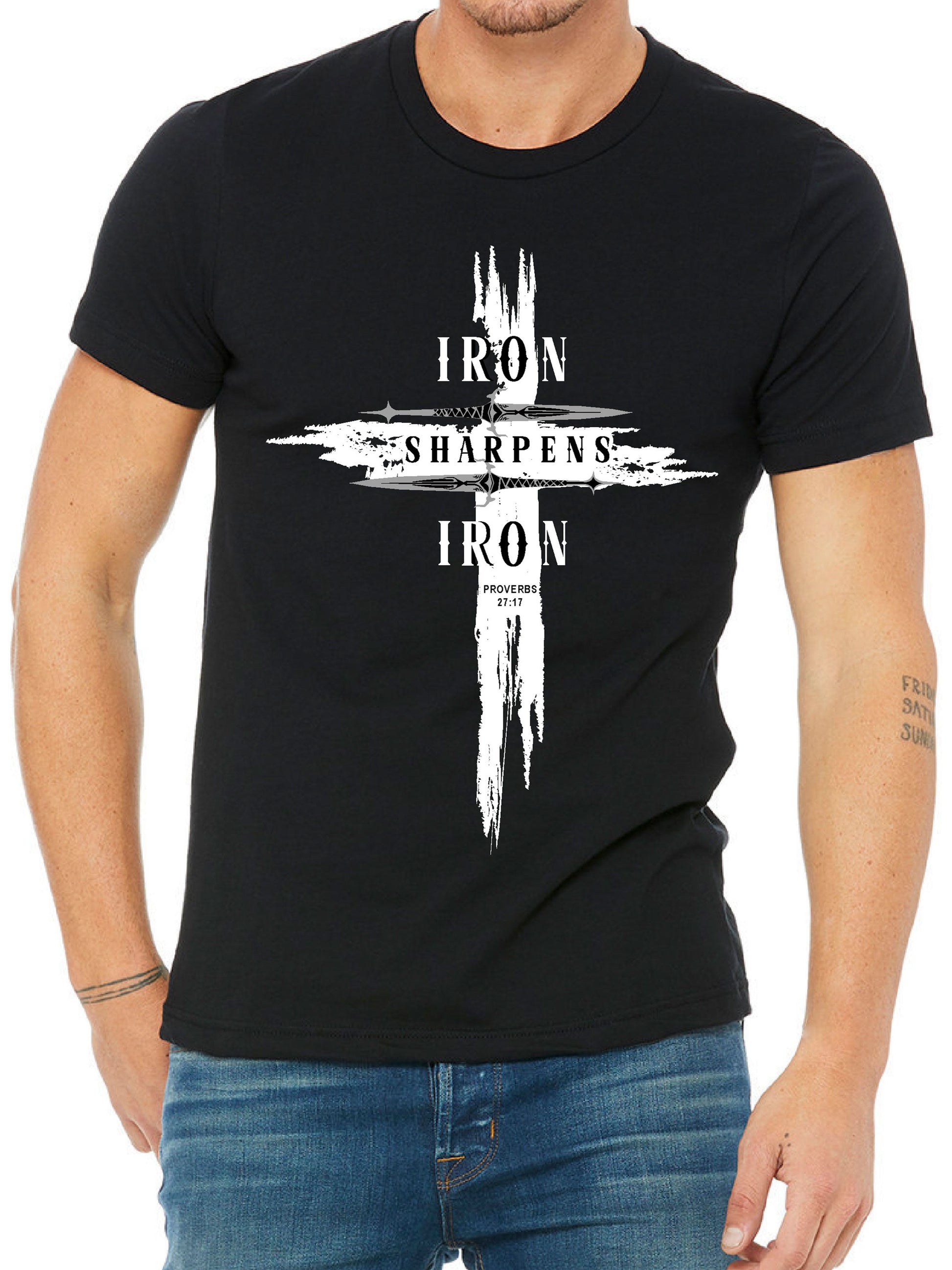 Man wearing a black T-shirt featuring a bold distressed cross design with the words ‘IRON SHARPENS IRON’ in white capital letters. Below the text, ‘Proverbs 27:17’ is printed. Styled with blue jeans.