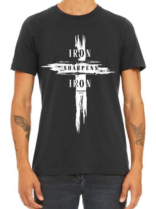 Man wearing a dark grey T-shirt featuring a bold distressed cross design with the words ‘IRON SHARPENS IRON’ in white capital letters. Below the text, ‘Proverbs 27:17’ is printed. Styled with black jeans.