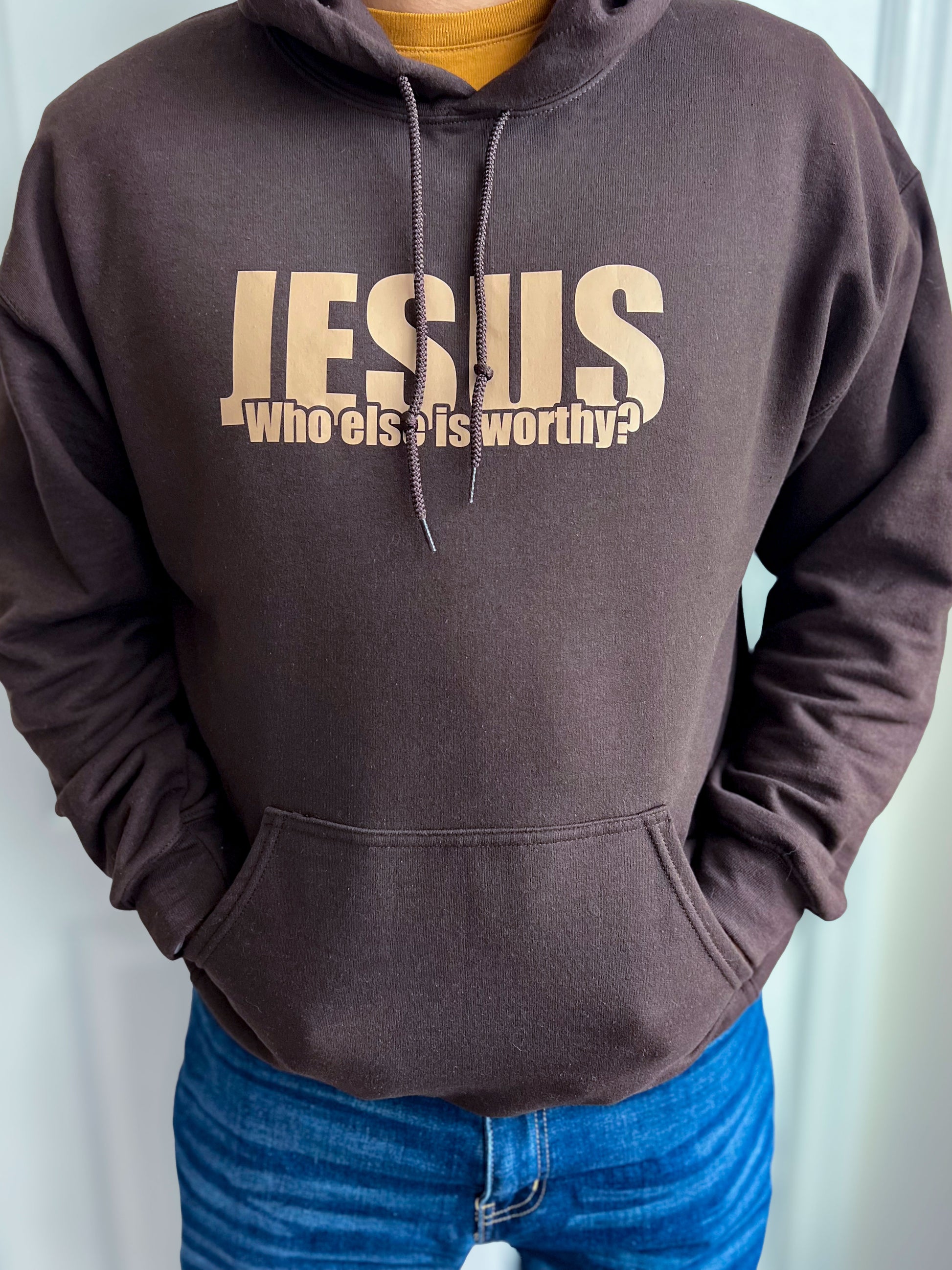 Man wearing a brown hoodie with bold beige text that reads ‘JESUS – Who else is worthy?’ Hands tucked in the front pocket, styled with blue jeans.