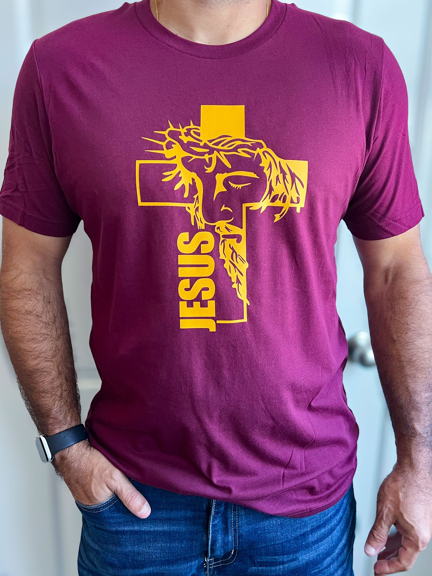 Man wearing a maroon T-shirt featuring a bold yellow design of Jesus’ face with a crown of thorns, integrated into a cross. The word ‘JESUS’ is printed vertically along the cross. Styled with blue jeans.