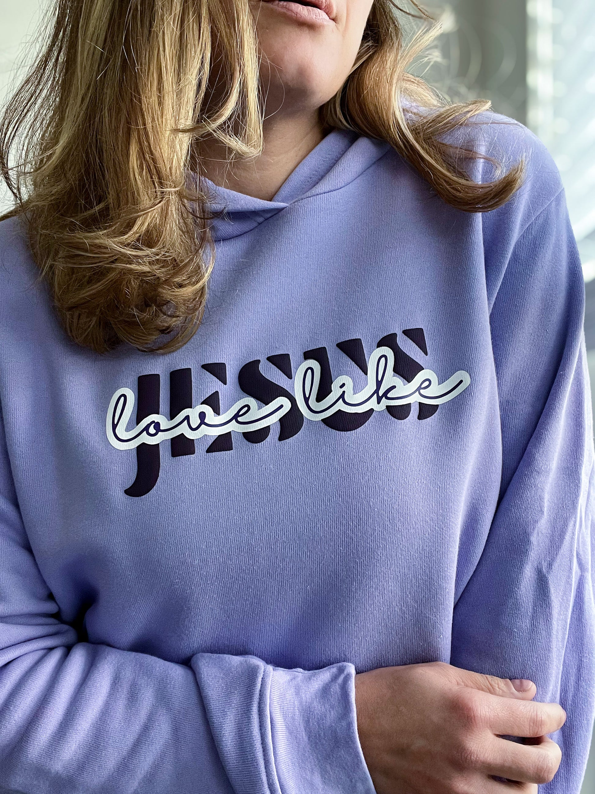 Close-up of a woman wearing a soft lavender hoodie with the phrase “Jesus Love Like” in a bold and stylish design. The word “Jesus” features a dark purple striped effect, while “love like” is in a white cursive font with a purple outline, creating a layered look. The hoodie is part of Spark of Faith’s handmade apparel collection, designed for faith-filled inspiration and everyday comfort.