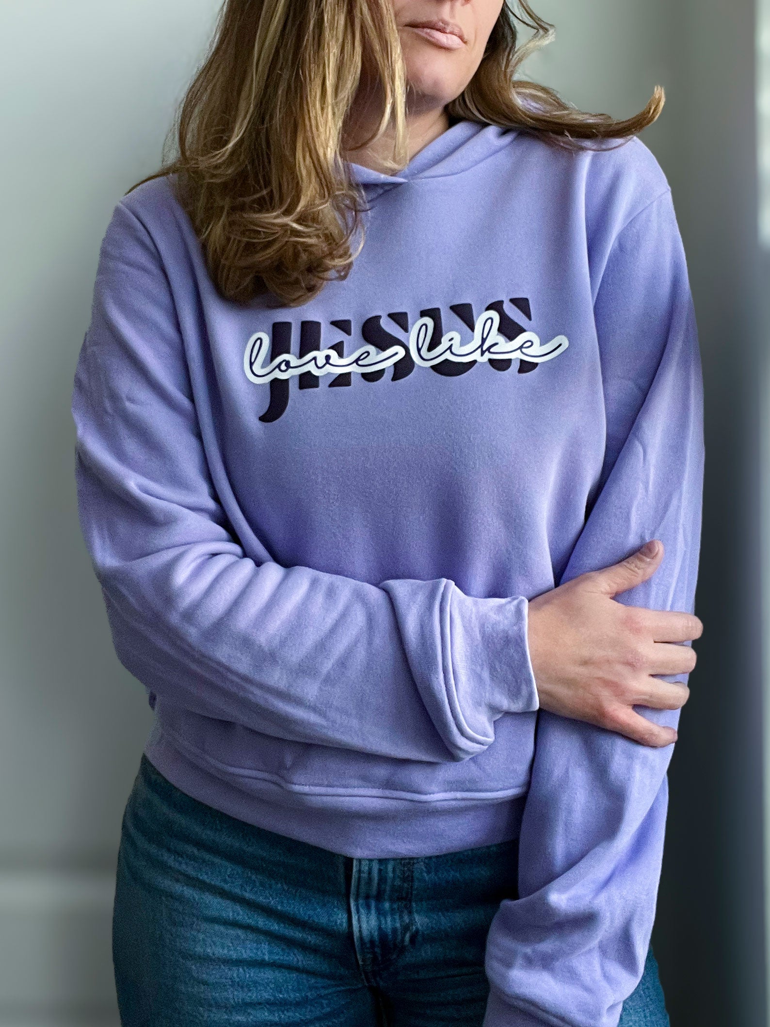 A woman wearing a soft lavender hoodie with the phrase “Jesus Love Like” in a bold and stylish design. The word “Jesus” is in dark purple with a striped effect, while “love like” is in a white cursive font over a purple outline. She pairs the hoodie with classic blue jeans for a cozy and faith-inspired look. Part of Spark of Faith’s handmade apparel collection.