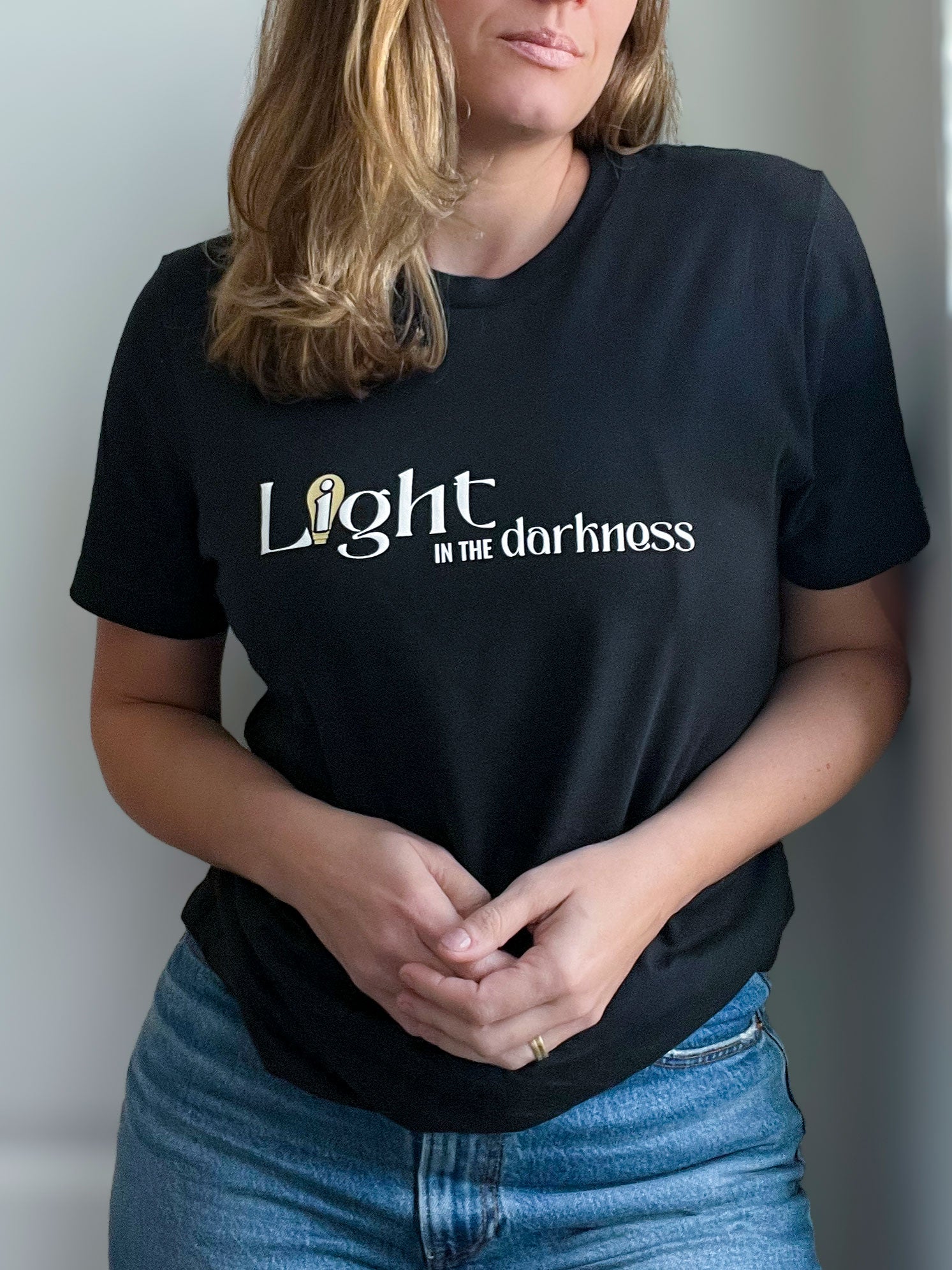 A woman wearing a vintage black T-shirt with the phrase “Light in the Darkness” in an elegant font. The word “Light” is accented with a stylized gold lightbulb, symbolizing faith and hope. She pairs the T-shirt with blue jeans for a casual yet meaningful look. Part of Spark of Faith’s handmade apparel collection.