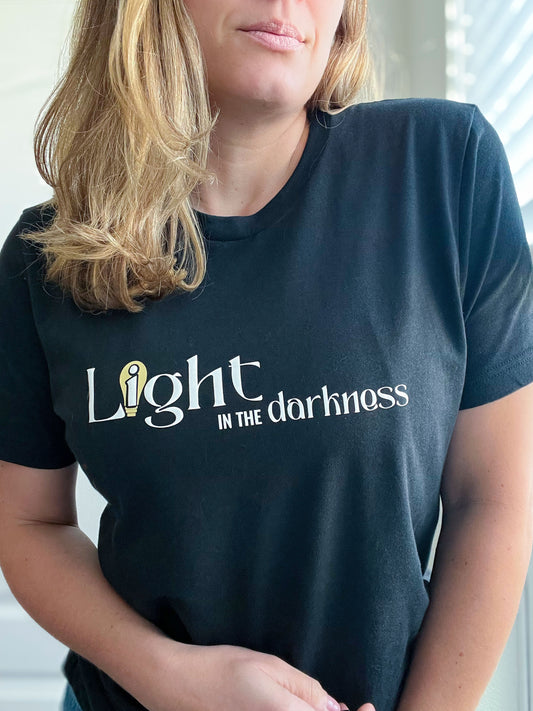 A woman wearing a vintage black T-shirt with the phrase “Light in the Darkness” in a mix of elegant and bold fonts. The word “Light” features a stylized lightbulb in gold, adding a symbolic touch to the faith-inspired design. The T-shirt is part of Spark of Faith’s handmade apparel collection, designed to inspire and uplift.