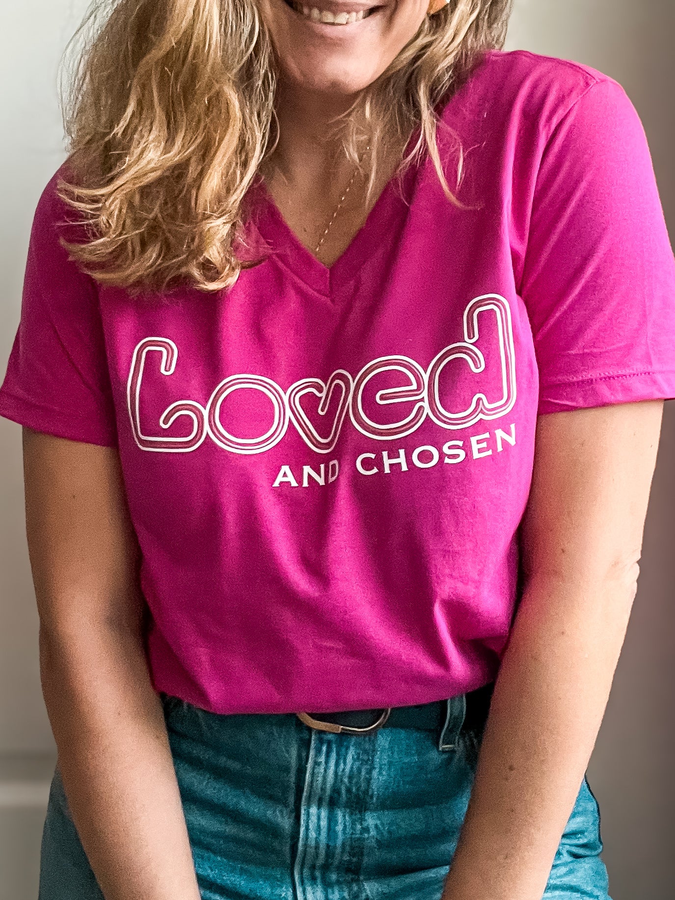 Woman wearing a vibrant pink V-neck T-shirt with bold white text that reads ‘Loved and Chosen.’ The word ‘Loved’ features a playful outlined. Styled with high-waisted blue jeans and a black belt.