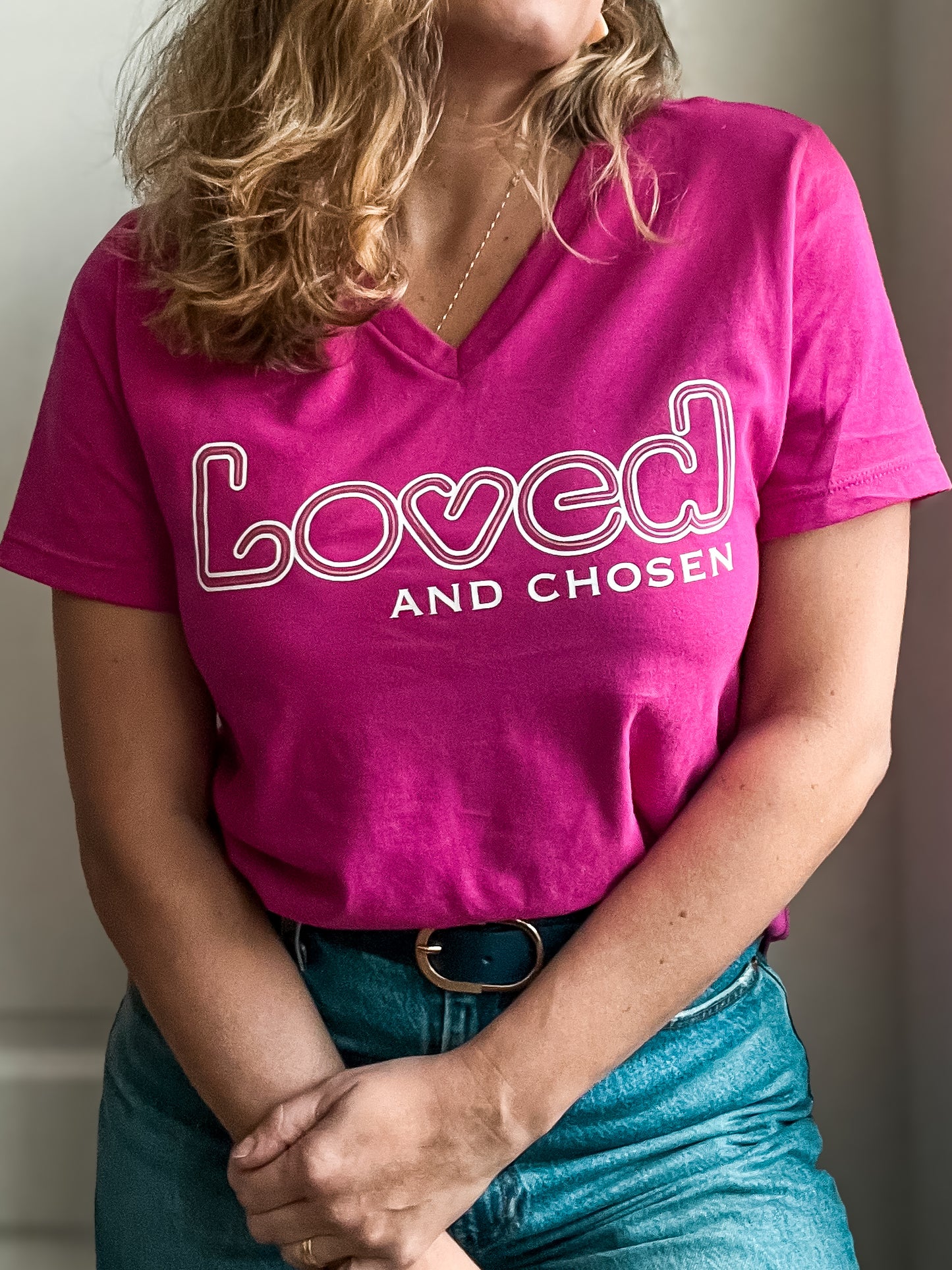 Woman wearing a bright pink V-neck T-shirt with ‘Loved and Chosen’ printed in white. The word ‘Loved’ features a playful outline. Styled with high-waisted blue jeans, a black belt with a gold buckle, and a delicate gold necklace.