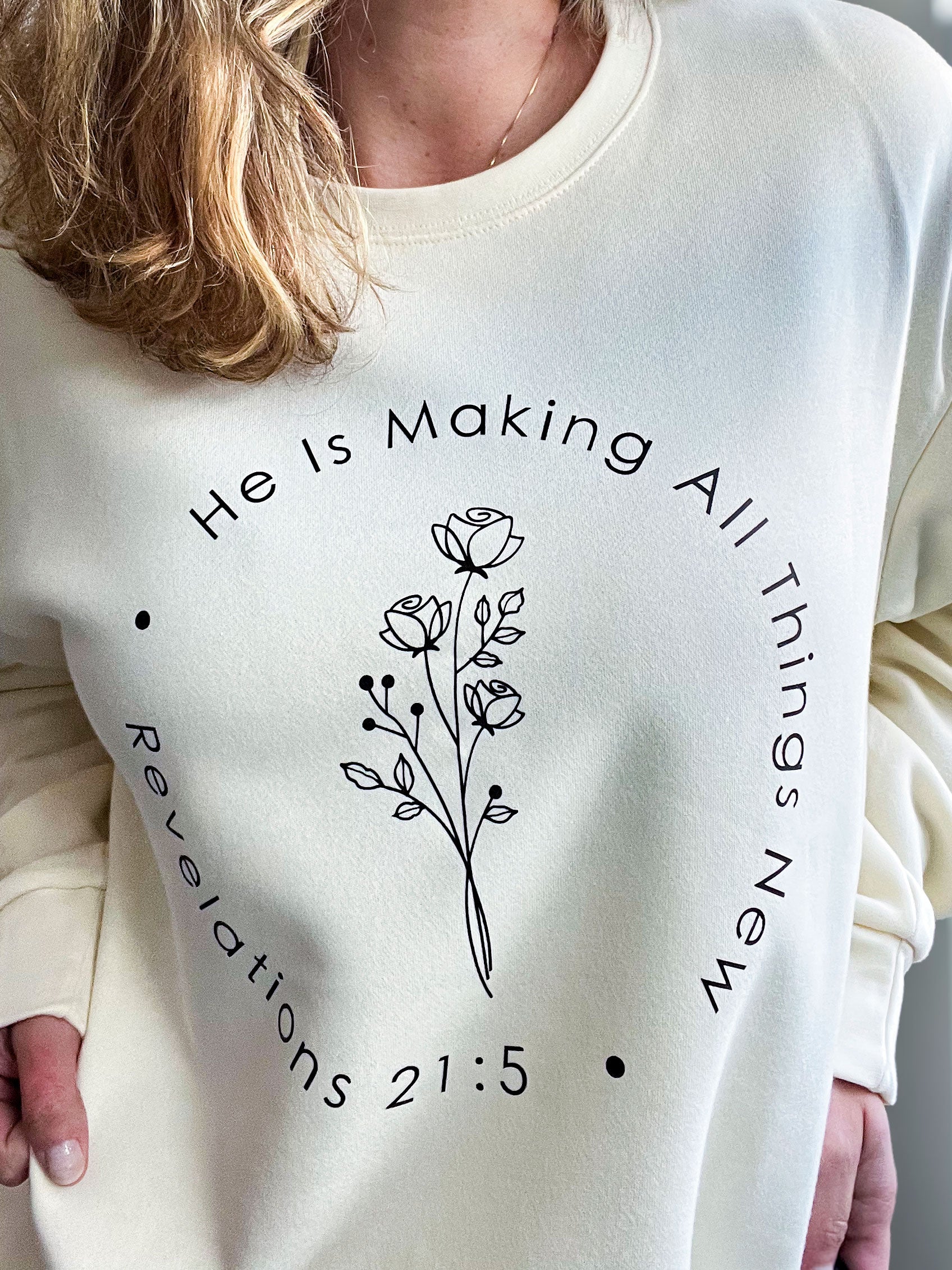 Woman wearing a cream oversized sweatshirt featuring a minimalist floral line-art design at the center. The text arcs around the design, reading ‘He Is Making All Things New’ with ‘Revelations 21:5’ printed below. Styled with a delicate gold necklace.