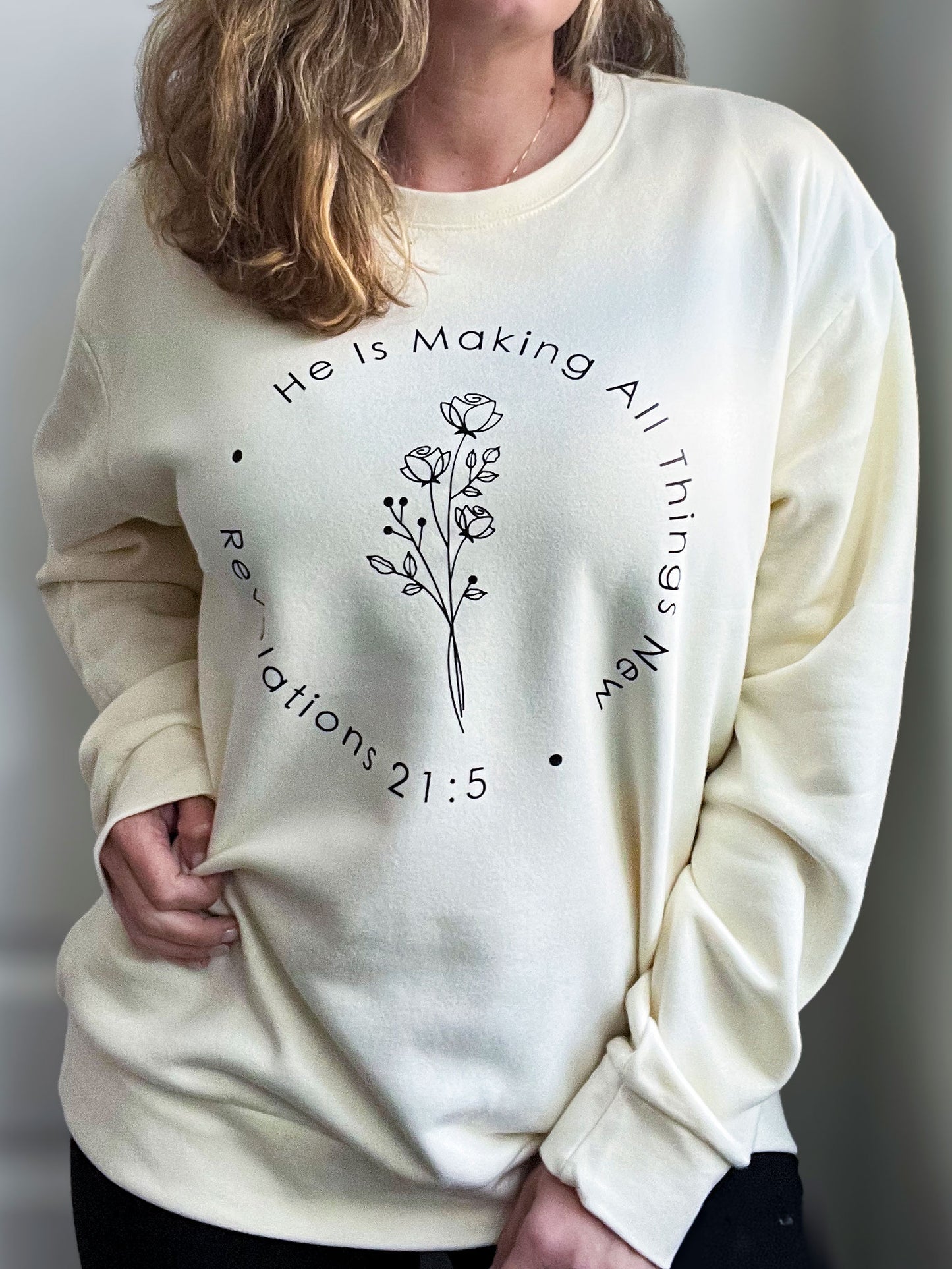 Woman wearing a cream oversized sweatshirt featuring a minimalist floral line-art design at the center. The text arcs around the design, reading ‘He Is Making All Things New’ with ‘Revelations 21:5’ printed below. Styled with a delicate gold necklace.