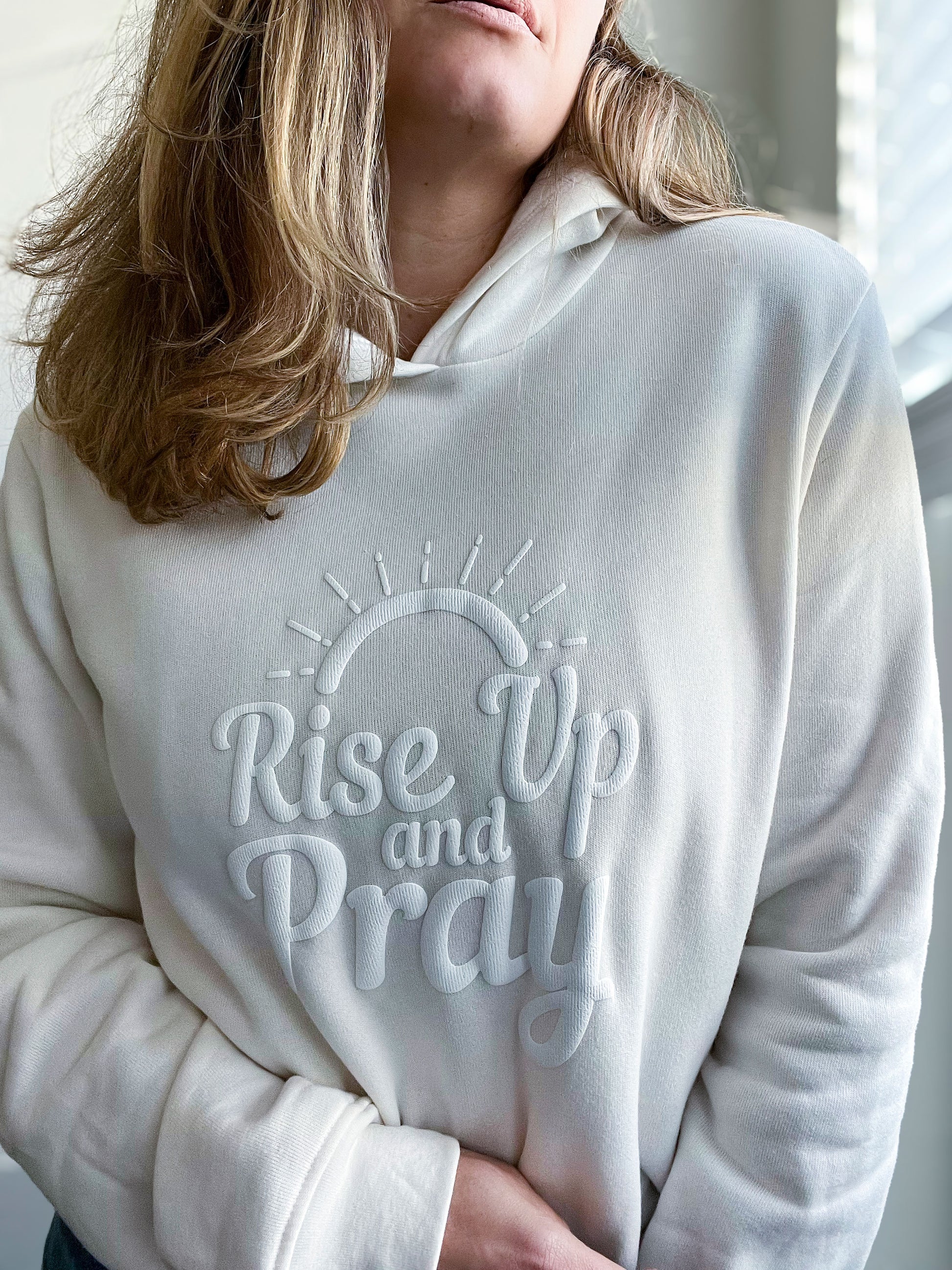 Close-up of a woman wearing a vintage white hoodie with the phrase “Rise Up and Pray” embossed in a raised, puff vinyl design. The text is accented with a minimalist sunrise illustration, creating a soft yet bold faith-inspired statement. The cozy hoodie is part of Spark of Faith’s handmade apparel collection.