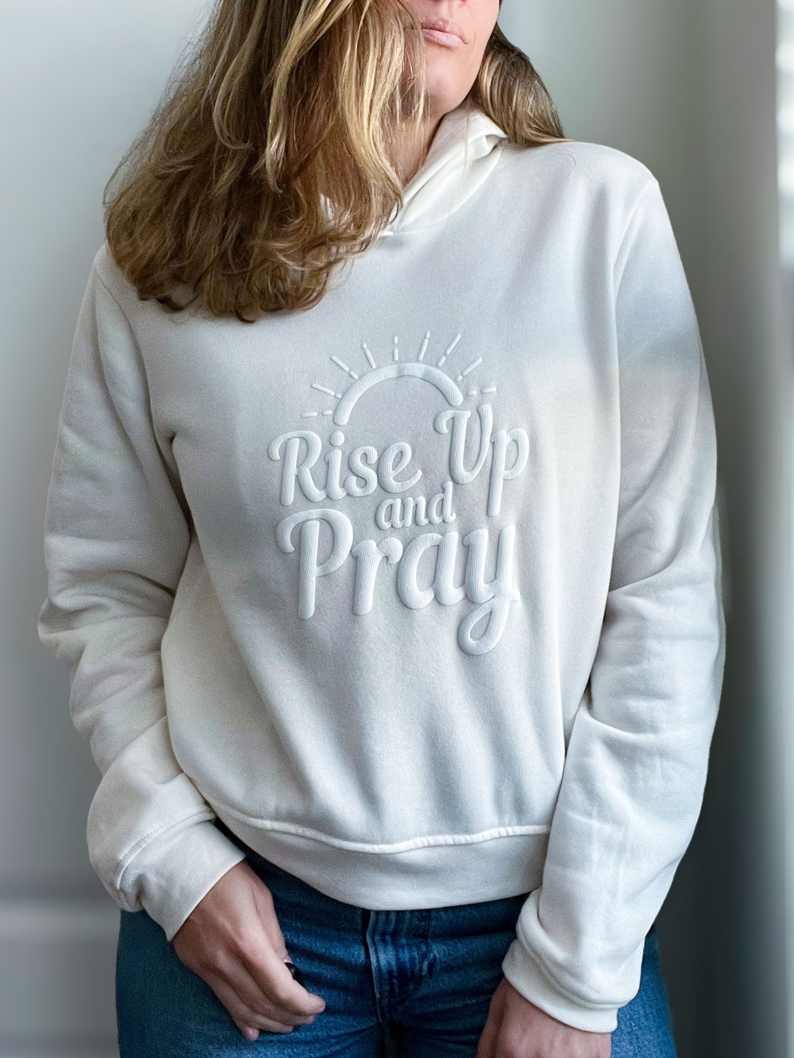 A woman wearing a soft, vintage white hoodie with the phrase “Rise Up and Pray” embossed in a raised, puff vinyl design. The text is styled with a minimalist sunrise illustration above it, creating a subtle yet bold faith-inspired statement. She pairs the hoodie with blue jeans for a cozy and uplifting look. Part of Spark of Faith’s handmade apparel collection.