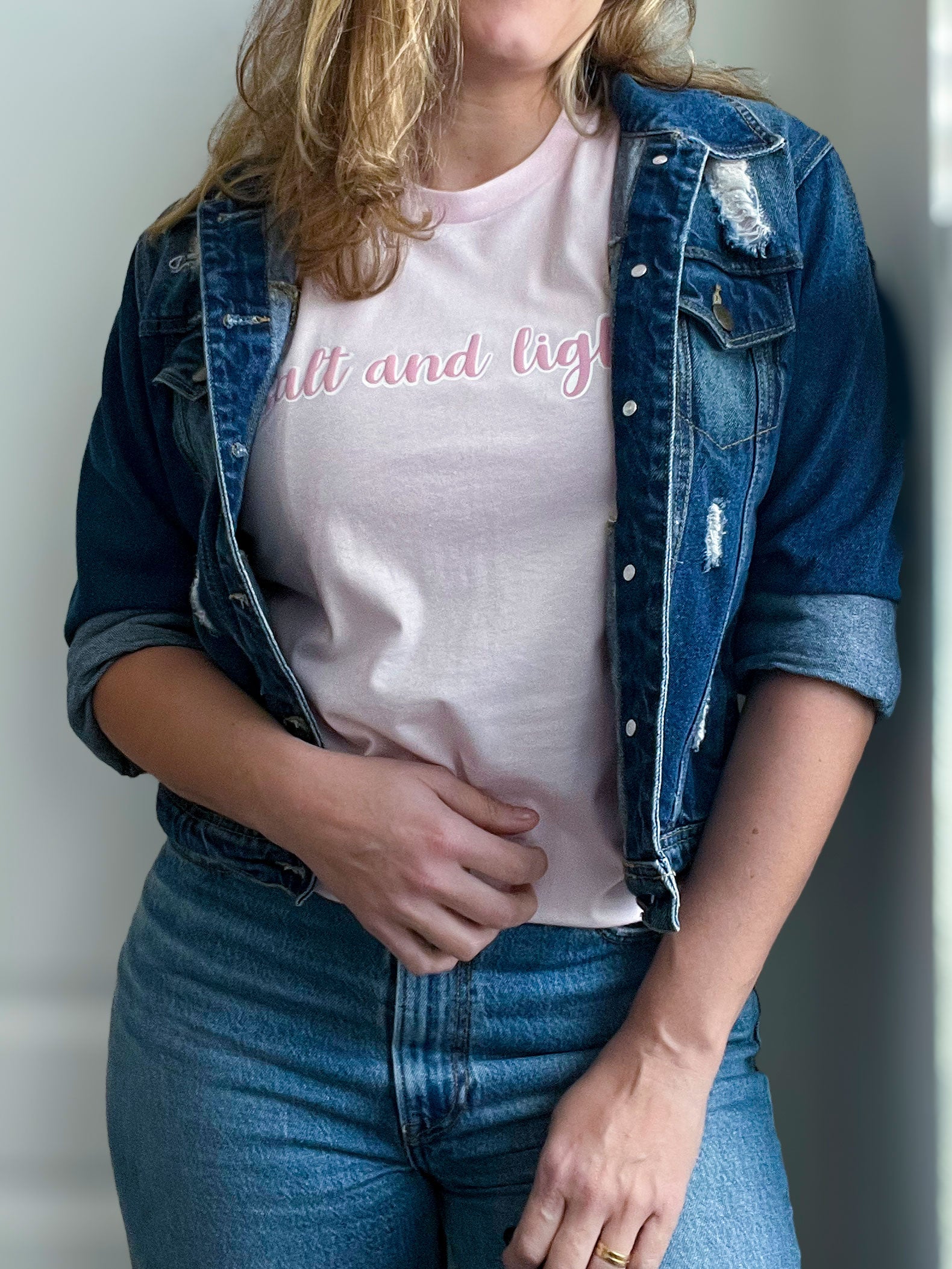 A woman wearing a light pink T-shirt with the phrase “salt and light” in a soft pink cursive font with a white outline. She pairs it with a distressed denim jacket and classic blue jeans for a stylish and casual faith-inspired look. Part of Spark of Faith’s handmade apparel collection.