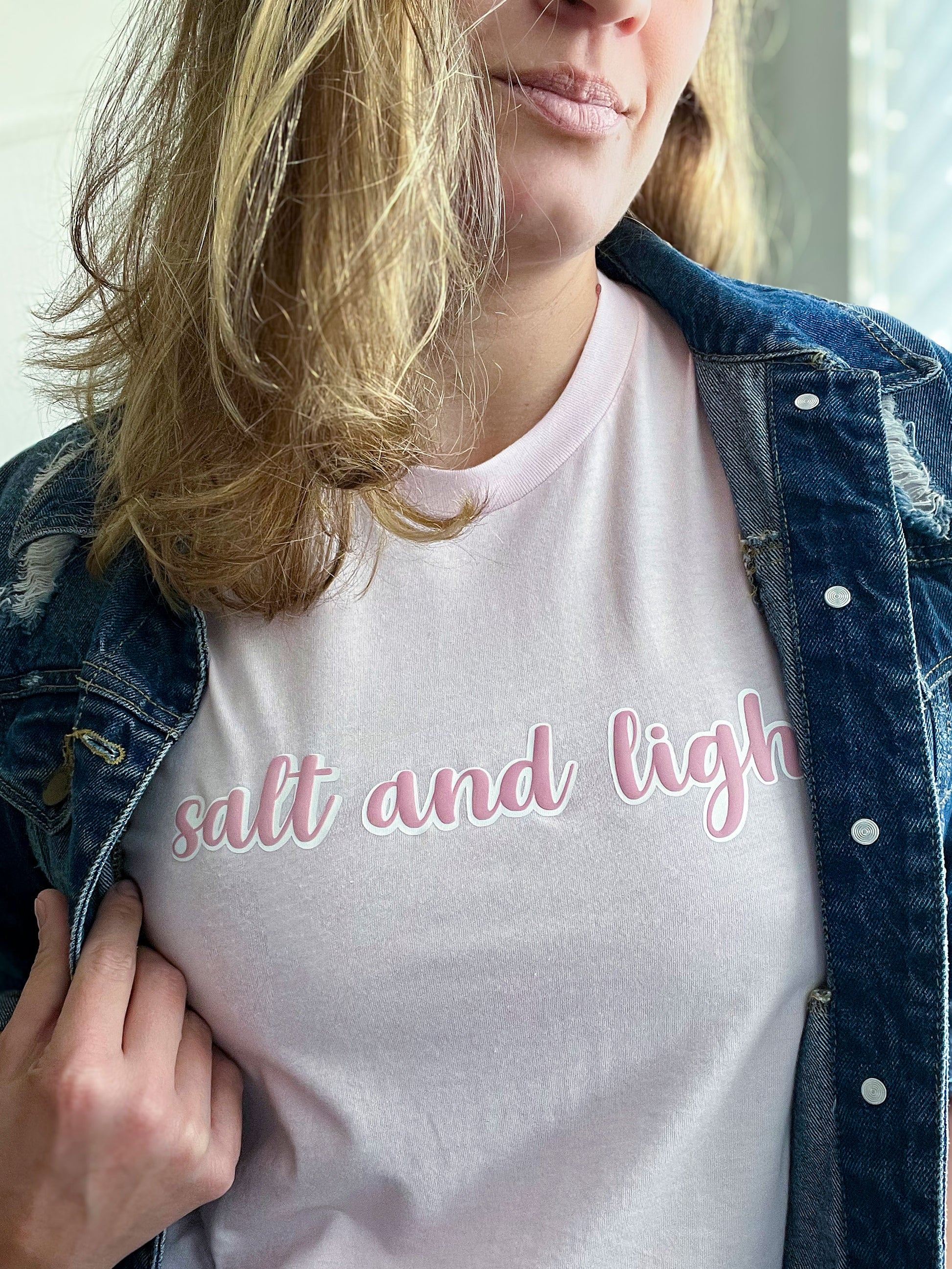 Close-up of a woman wearing a light pink T-shirt with the phrase “salt and light” in a soft pink cursive font with a white outline. She pairs it with a distressed denim jacket, slightly pulled open to reveal the faith-inspired design. Part of Spark of Faith’s handmade apparel collection.