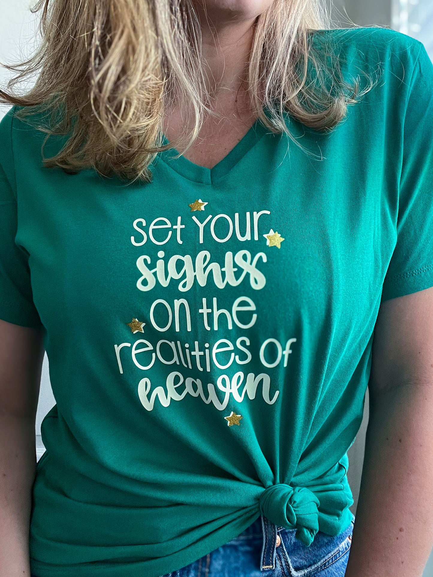 Close-up of a woman wearing a green V-neck T-shirt with the phrase “Set your sights on the realities of heaven” in beige lettering. The design is accented with small gold stars, adding a sparkling touch. The T-shirt is tied in a front knot, creating a stylish and casual look. Part of Spark of Faith’s handmade apparel collection.