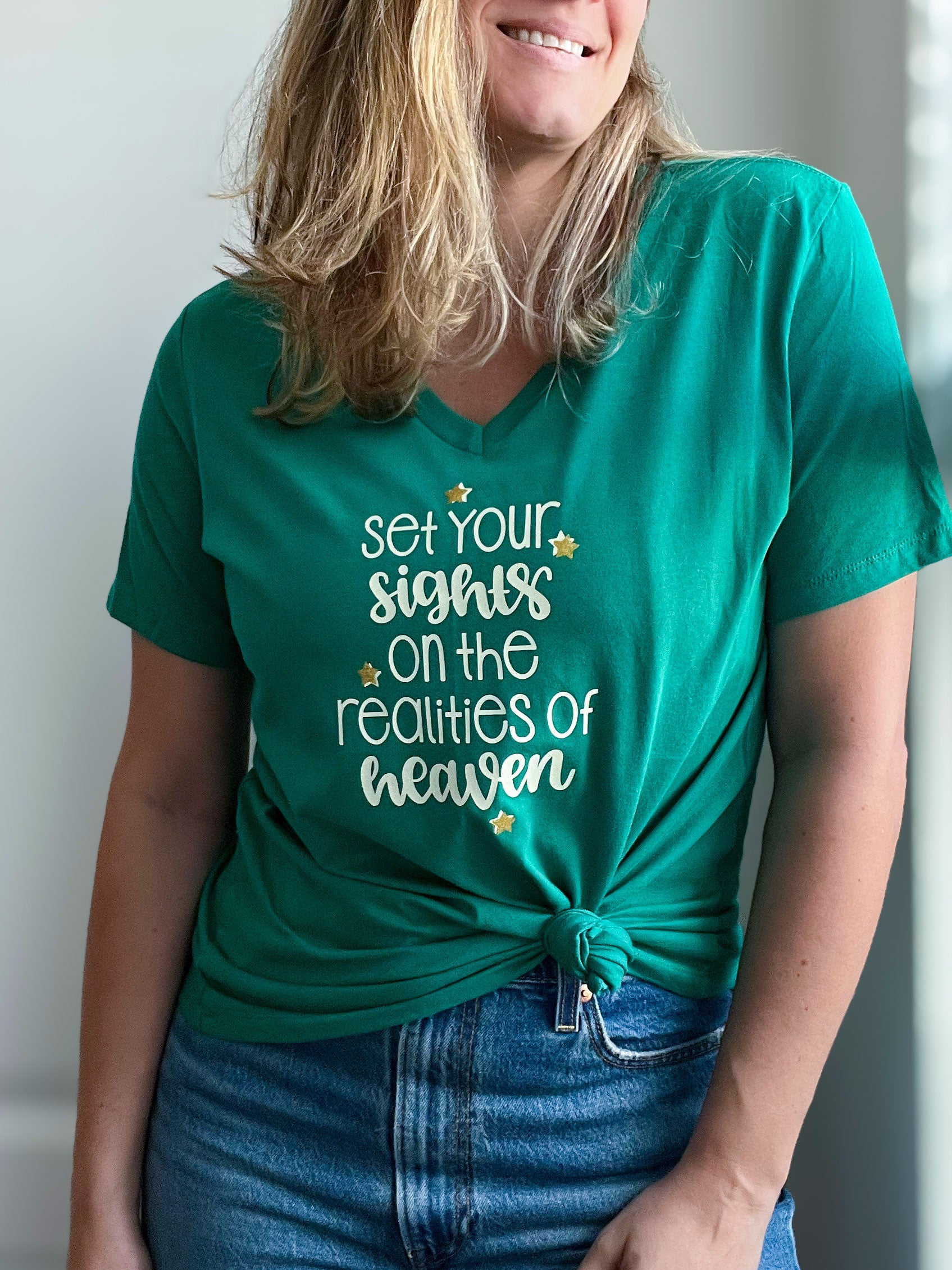 A woman wearing a vibrant green V-neck T-shirt with the phrase “Set your sights on the realities of heaven” in a mix of bold and cursive beige lettering. Small gold stars accent the design, adding a sparkling touch. She has the shirt stylishly knotted at the front and pairs it with blue jeans for a casual, faith-filled look. Part of Spark of Faith’s handmade apparel collection.