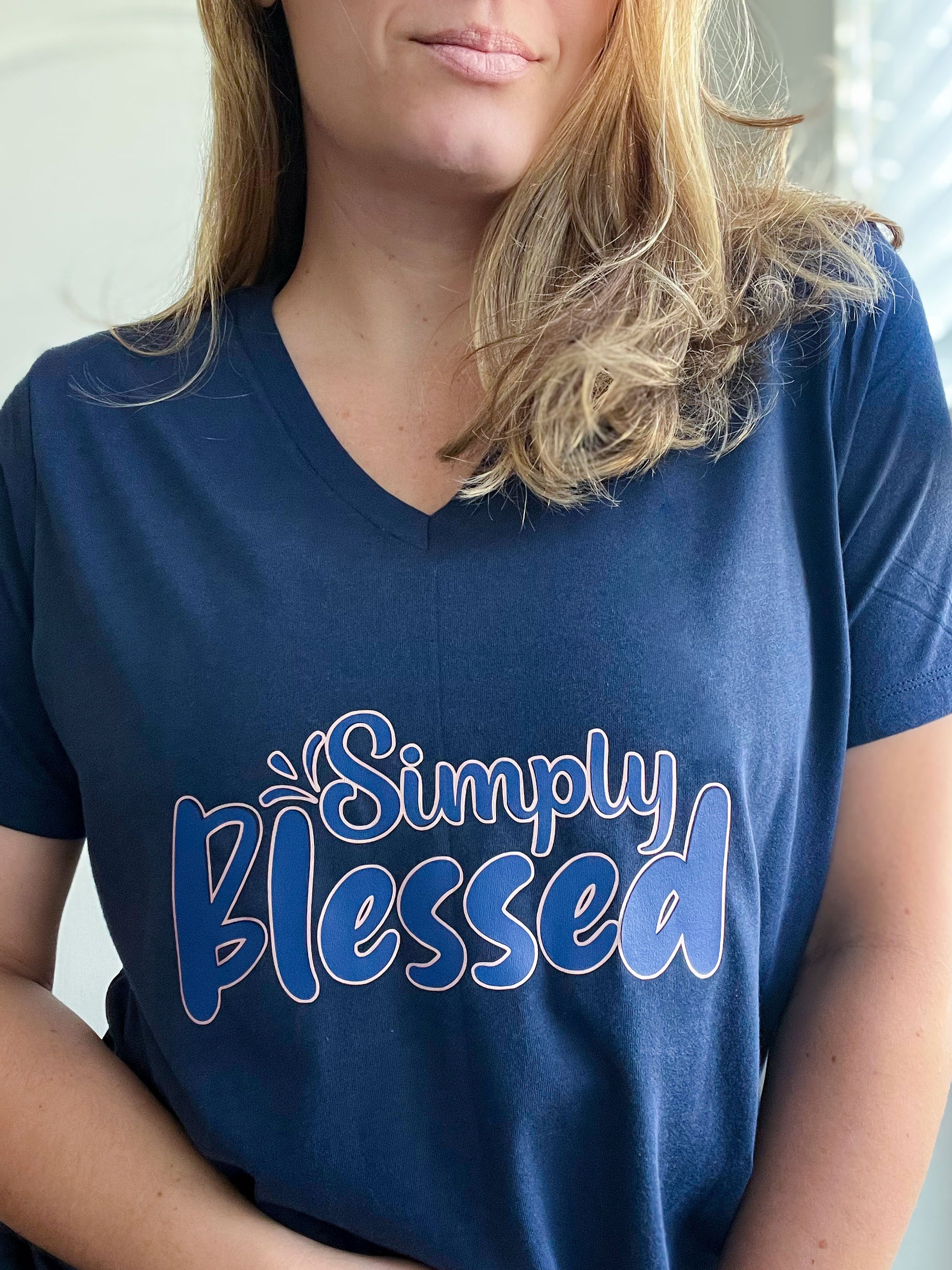 Close-up of a woman wearing a navy blue V-neck T-shirt with the phrase “Simply Blessed” in a playful font. The text design features a blue text with a light pink outline, adding a soft contrast to the dark fabric. The T-shirt is part of Spark of Faith’s faith-inspired apparel collection.