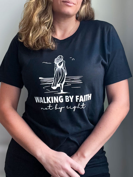 Woman wearing a vintage black T-shirt featuring a white line-art illustration of a woman walking along the shore. Below the design, the text reads ‘WALKING BY FAITH’ in bold uppercase letters, followed by ‘not by sight’ in elegant script. Styled with a delicate gold necklace.