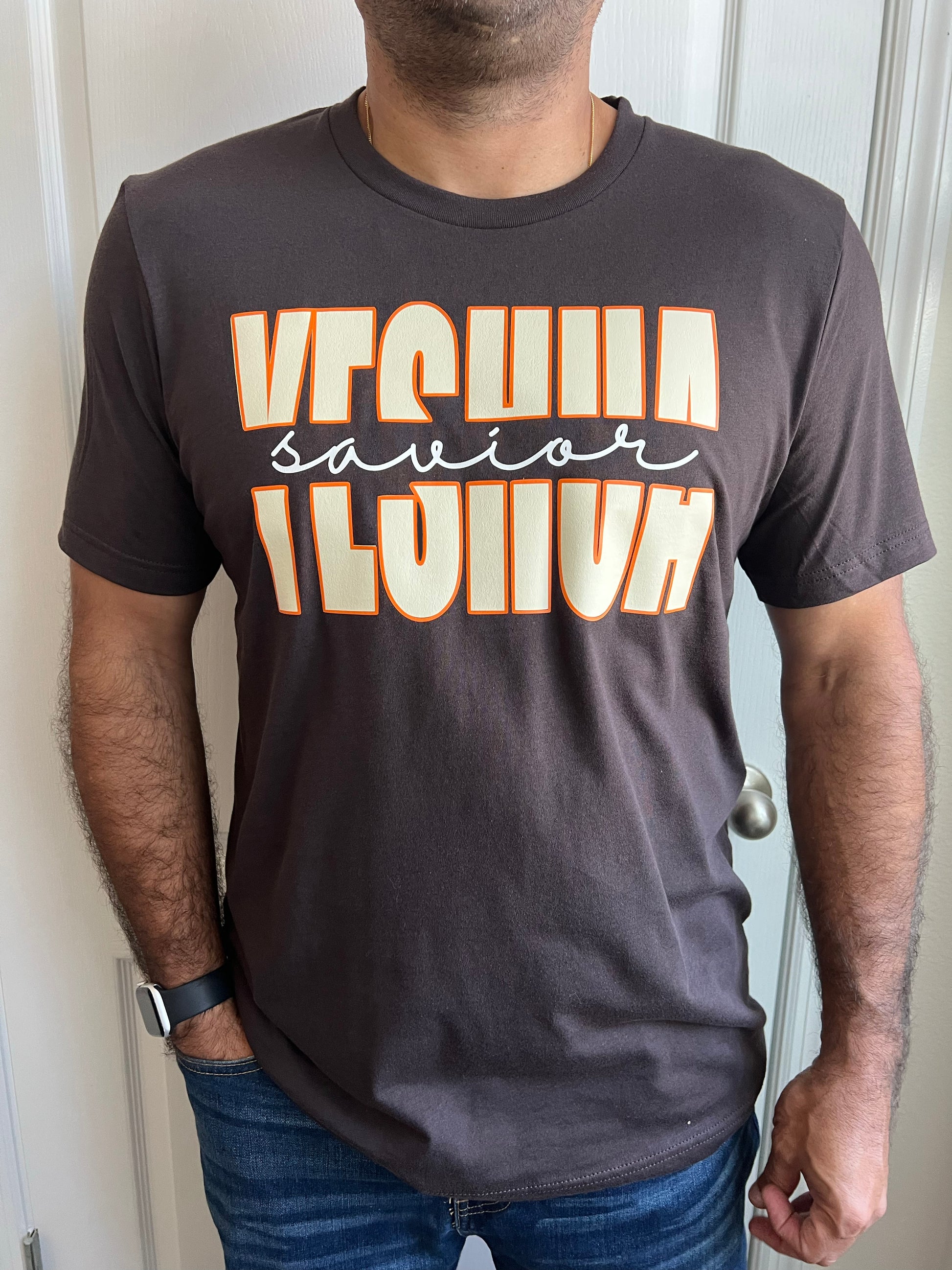 Man wearing a dark brown T-shirt with bold cream and orange text reading ‘YESHUA’ with ‘savior’ in elegant white script between the words. Styled with blue jeans.